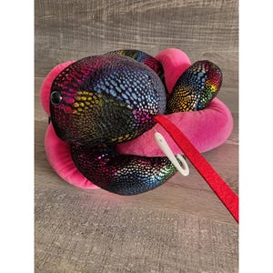 Op de afbeelding: Een pluche speelgoedslang met een glinsterend, veelkleurig schubbenpatroon in blauw, paars en goud. De slang is omgeven door een roze, ringvormig kussen. Een rode riem met een witte clip is bevestigd.