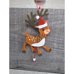 May include: A festive Christmas ornament featuring a reindeer design. The reindeer is brown with white spots, wearing a red Santa hat and a green and red striped scarf. It has brown antlers and a red nose. The ornament hangs from a red string.