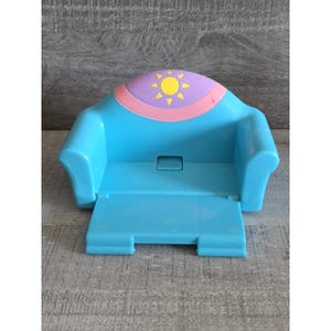 May include: A light blue plastic toy sofa with a fold-out footrest. The back of the sofa has a purple and pink design with a yellow sun. The toy sofa is designed for dolls or small figures.