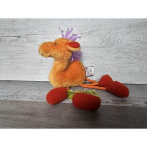 May include: Orange plush camel toy with purple hair and red feet. The camel has a tag on its back that says "Cuddlekins".