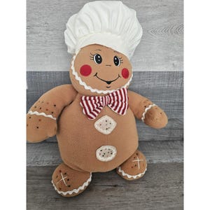 May include: A gingerbread man plush toy wearing a white chef's hat and a red and white striped bow tie. The toy has a brown fuzzy body with white icing details and a smiling face.