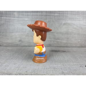 May include: A Little People Woody figure, featuring a brown cowboy hat, yellow shirt, red neckerchief, and blue pants. The figure has brown hair and a tan face. The base of the figure is brown and has the words "Little People" printed on it.