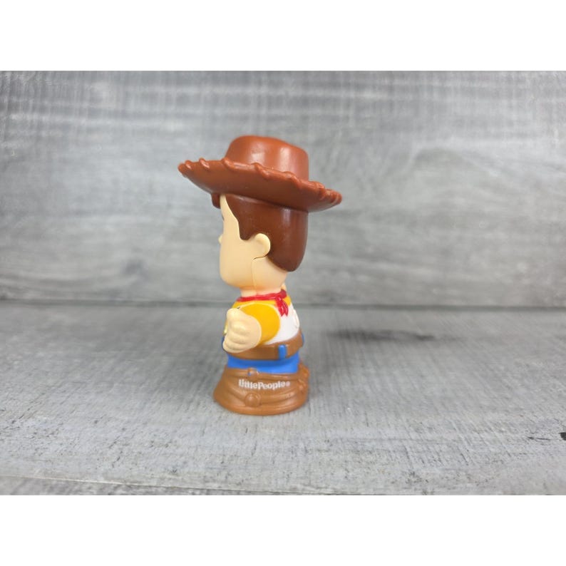 May include: A Little People Woody figure, featuring a brown cowboy hat, yellow shirt, red neckerchief, and blue pants. The figure has brown hair and a tan face. The base of the figure is brown and has the words "Little People" printed on it.