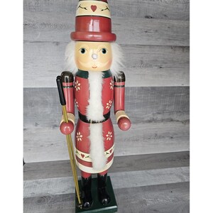 Könnte beinhalten: Eine hölzerne Nussknackerfigur, die ein rot-weißes Outfit mit weißem Pelzbesatz trägt. Der Nussknacker hält zwei hölzerne Skistöcke und steht auf einem grünen Sockel.