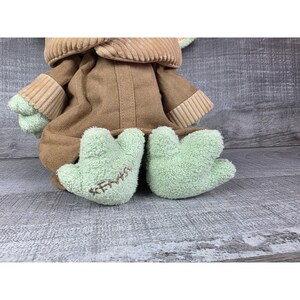 May include: A plush doll of a character from a popular science fiction franchise. The doll has a brown coat, tan corduroy collar, and light green feet. The feet have the word "KENYA" embroidered on them.