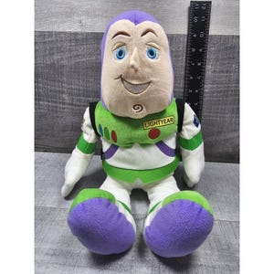 May include: A plush Buzz Lightyear doll, with a purple head, blue eyes, and a smiling expression. The doll wears a green and white spacesuit with the word "LIGHTYEAR" on the chest. The doll is approximately 25 cm tall.