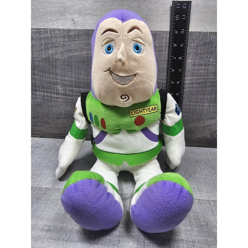 May include: A plush Buzz Lightyear doll, with a purple head, blue eyes, and a smiling expression. The doll wears a green and white spacesuit with the word "LIGHTYEAR" on the chest. The doll is approximately 25 cm tall.