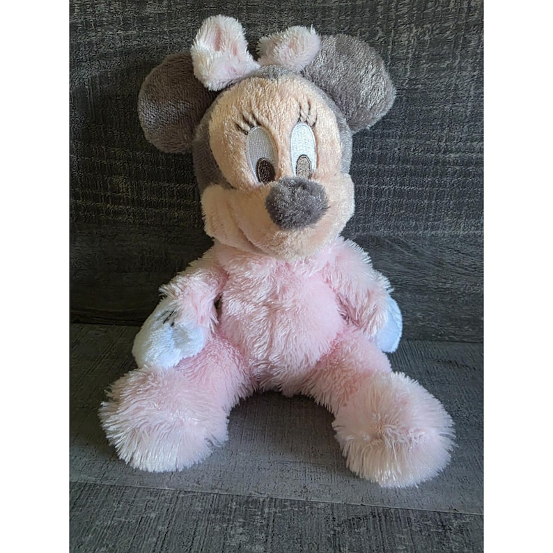 May include: A plush Minnie Mouse doll in a seated position. The doll features a pink body, gray ears, and a light pink face with embroidered details. The doll's hands and feet have white and light blue accents.