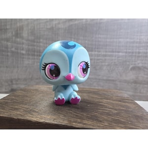 May include: A light blue figurine of a cartoon bird with large eyes, eyelashes, and a pink beak. The bird has a blue head with a darker blue swirl design. The eyes are pink and black with white star accents. The figurine is sitting on a wooden surface.