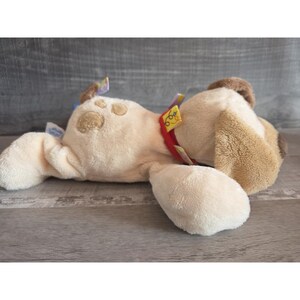 Puede incluir: Un juguete de peluche de perro en crema y marr&oacute;n, tumbado de lado. El perro tiene un collar rojo y detalles de huellas de patas. Hay etiquetas coloridas adjuntas a la oreja y al collar. El juguete est&aacute; sobre una superficie de madera gris.