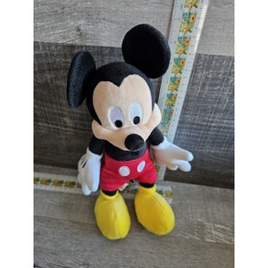 Puede incluir: Un juguete de peluche de Mickey Mouse con orejas negras, cara negra, ojos blancos, camisa roja y blanca y zapatos amarillos.