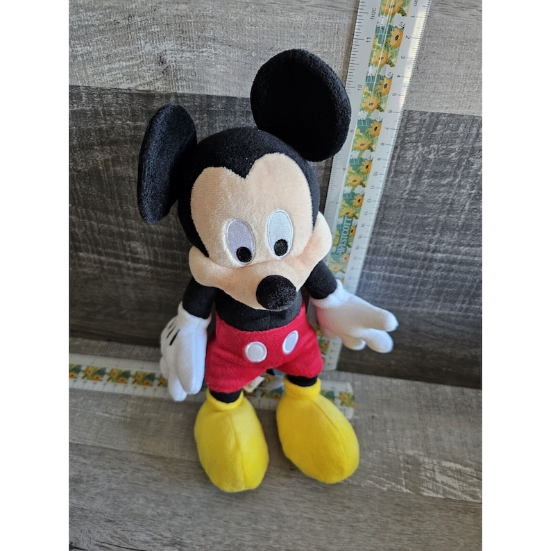 Puede incluir: Un juguete de peluche de Mickey Mouse con orejas negras, cara negra, ojos blancos, camisa roja y blanca y zapatos amarillos.