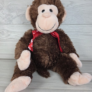 May include: A plush monkey toy with dark brown shaggy fur, a light beige face, hands, and feet. The monkey features black embroidered eyes and a stitched smile, and it is wearing a red satin ribbon tied around its neck with small white heart patterns.