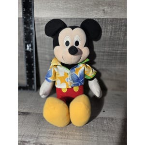 Puede incluir: Mu&ntilde;eco de peluche de Mickey Mouse con una camisa hawaiana con estampados florales amarillos, azules y verdes. Mickey tiene orejas negras, una nariz negra y zapatos amarillos. Lleva pantalones cortos rojos.