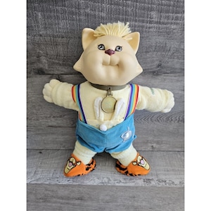 Peut inclure: Poupée chat en peluche avec un visage crème, des cheveux jaunes et une chemise à rayures arc-en-ciel. Elle porte un short en velours côtelé bleu, des chaussures à rayures tigrées orange et noires et un collier doré avec un pendentif.