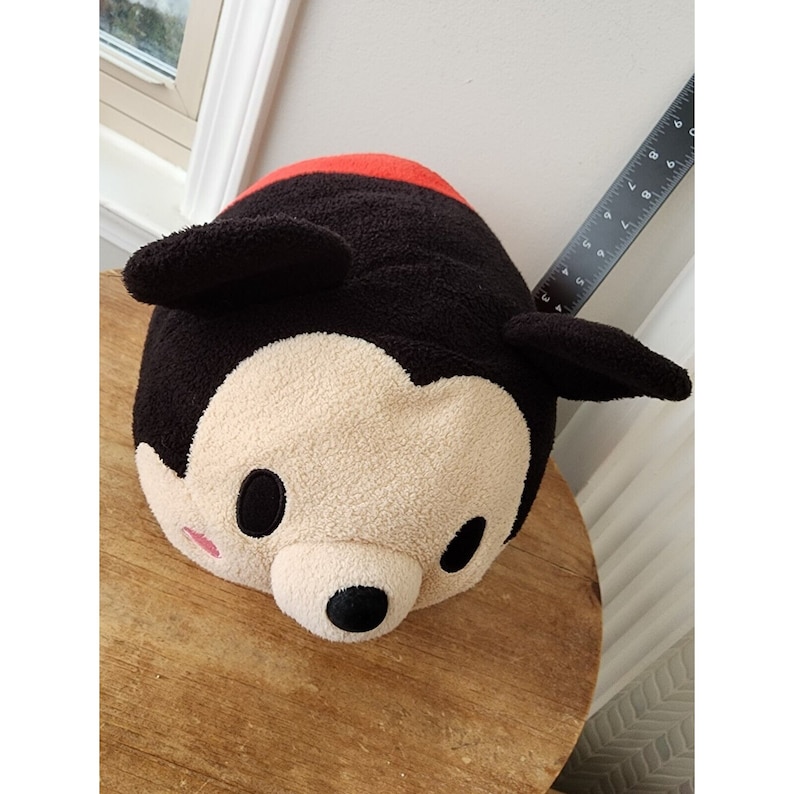 May include: A plush Mickey Mouse toy with black ears, a red nose, and black eyes. The toy is made of soft, fuzzy material and is shaped like a round pillow.