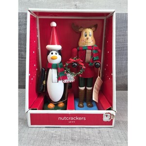 May include: A set of two Christmas nutcrackers in a box. One is a penguin wearing a red and white hat and a green and red scarf. The other is a reindeer with a red outfit and a green and red scarf. The box reads "nutcrackers set of 2".