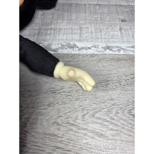 May include: Close-up of a plush toy arm and hand. The arm is covered in black fabric, and the hand is a light beige color. The hand is in a relaxed, open position.
