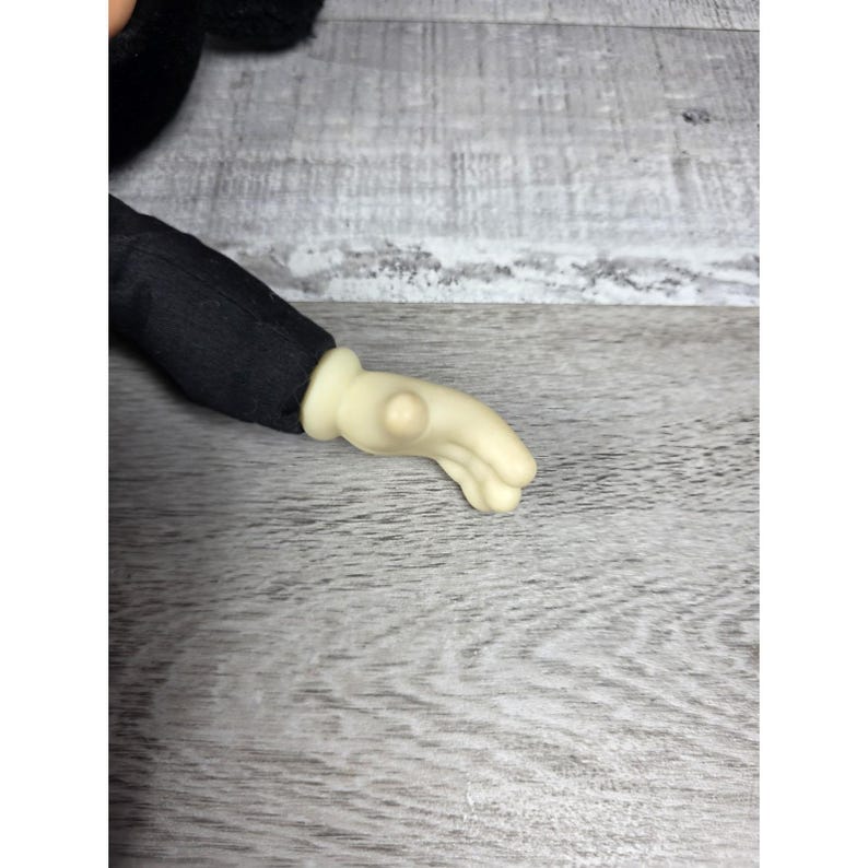 May include: Close-up of a plush toy arm and hand. The arm is covered in black fabric, and the hand is a light beige color. The hand is in a relaxed, open position.