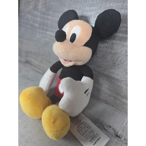 May include: A plush Mickey Mouse toy. The toy features a black body, ears, and tail, with a tan face and white gloves. It has yellow feet and red shorts. The toy is sitting on a gray surface.