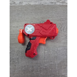 Può includere: Una pistola giocattolo rossa e arancione con grilletto, impugnatura e un emblema circolare con una "X" bianca su sfondo blu. La pistola giocattolo ha un design strutturato ed è realizzata in plastica.