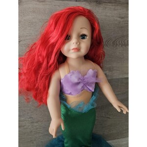 Puede incluir: Una mu&ntilde;eca de pelo rojo con un disfraz de sirena morado y verde. La mu&ntilde;eca tiene ojos azules y un tono de piel claro.
