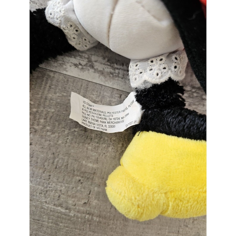 K&ouml;nnte beinhalten: Ein schwarzes und gelbes Pl&uuml;schtier mit einem wei&szlig;en Kragen und einer wei&szlig;en Spitzenborte. Das Spielzeug hat ein Etikett mit der Aufschrift "Disney, All New Materials, Polyester Fibres, Polythylene Pellets, Reg. No. PA 125400, OH 18724, MB 3000, Disney Theme Park Merchandise, Lake Buena Vista, FL 32830, Product of Vietnam, DVT027JH".