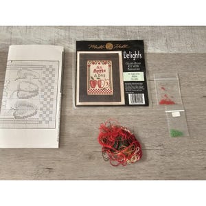 Può includere: Un kit di ricamo a punto croce con un design di mela rossa e verde. Il kit include perline di vetro, filo e uno schema. La confezione presenta il testo "An Apple A Day" e "Mile Hill Delights".