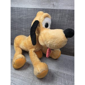 Pode incluir: Um brinquedo de pelúcia do Pluto, um cão amarelo com orelhas pretas e um nariz preto. O brinquedo tem uma língua rosa saindo e olhos grandes e redondos. O cão está sentado com as pernas estendidas.