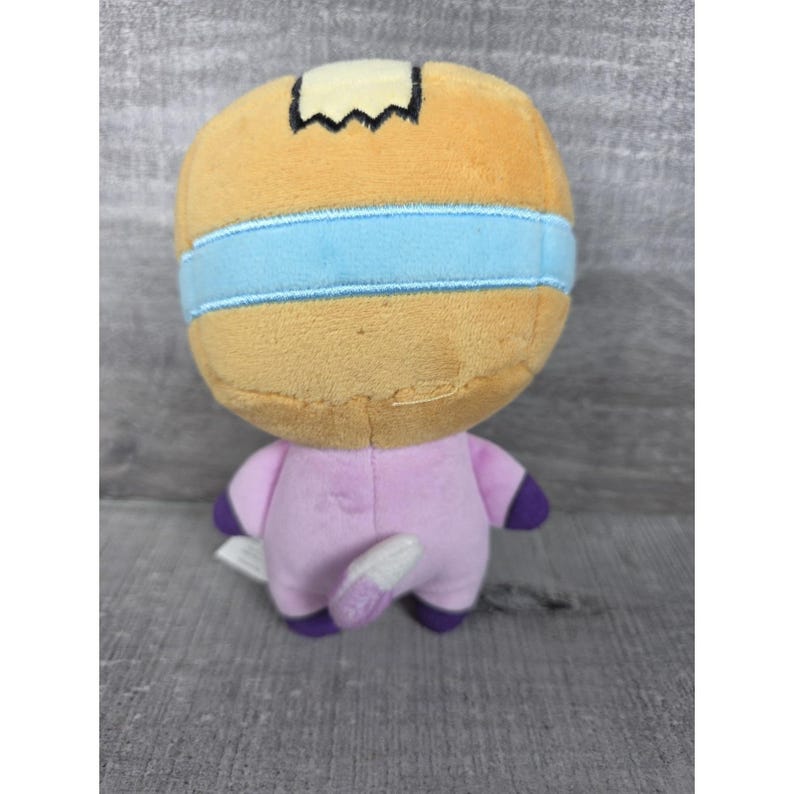 May include: A plush toy featuring a tan, rounded head with a light blue horizontal band and a small black patch on top. The body is light pink with dark purple accents on the feet and a small, light-colored tail extending from the back.