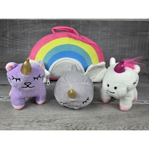 Könnte beinhalten: Eine Sammlung von Plüschtieren, darunter eine lila Katze mit goldenem Horn, ein grauer Narwal mit goldenem Horn und ein weißes Einhorn mit rosa Horn. Im Hintergrund ist eine regenbogenförmige Tasche zu sehen.