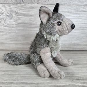 May include: A plush wolf toy featuring a grey and white textured coat, pointed ears with black tips, dark brown eyes, and a black nose. The stuffed animal is depicted in a seated position with a long, bushy tail extending behind its body.