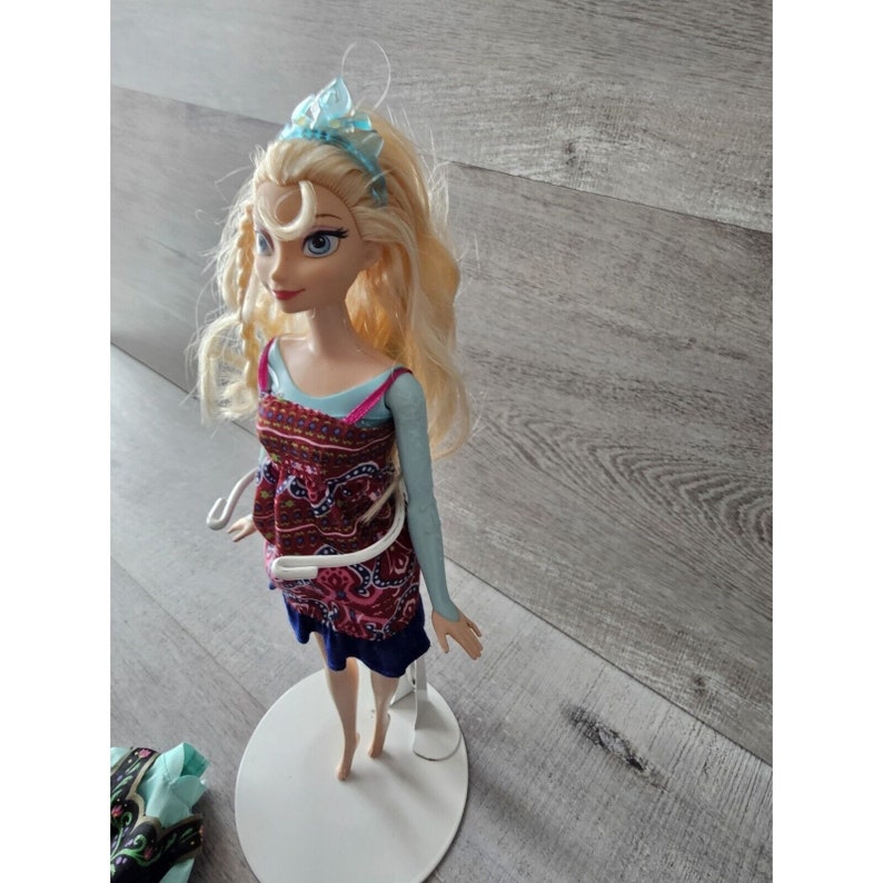 Puede incluir: Una mu&ntilde;eca rubia con una corona azul, vestida con un vestido estampado en azul y rojo, de pie sobre un soporte blanco.