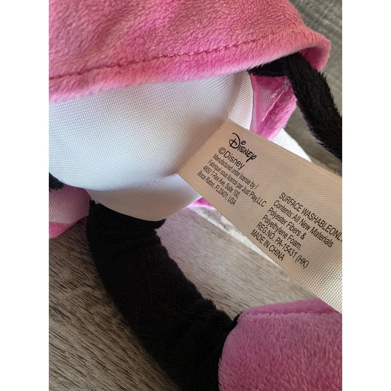 May include: A plush toy featuring a pink bow and dress, black ears, and a white face. The toy has a tag that reads "Disney" and "SURFACE WASHABLEONL". The toy is made of polyester fibers and polyethylene foam.
