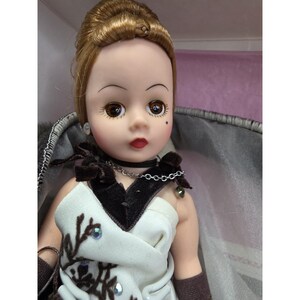 May include: A vintage doll with brown eyes, red lips, and a beauty spot. The doll has light brown hair styled up and wears a white and brown dress with embellishments. It has a black choker necklace and a silver chain.