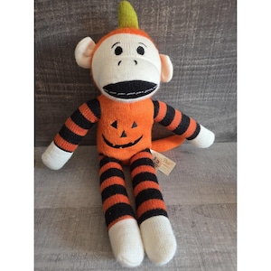 May include: A Halloween-themed sock monkey doll. The monkey has a white face, ears, and hands, with a black mouth and eyes. The body is orange with a jack-o'-lantern face and black and orange striped arms and legs. A green stem tops the head.