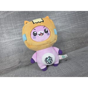 May include: A plush toy featuring a purple character wearing a tan, box-shaped headpiece with small ears. The character has large black eyes, a small mouth, and a white circular patch on its chest with a stylized letter F inside a jagged border.