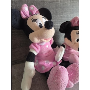 May include: Two Minnie Mouse plush toys. The plush toys have black ears, a beige face, and a black nose. They are wearing pink dresses with white polka dots and pink bows. The plush toys have pink sparkly shoes.