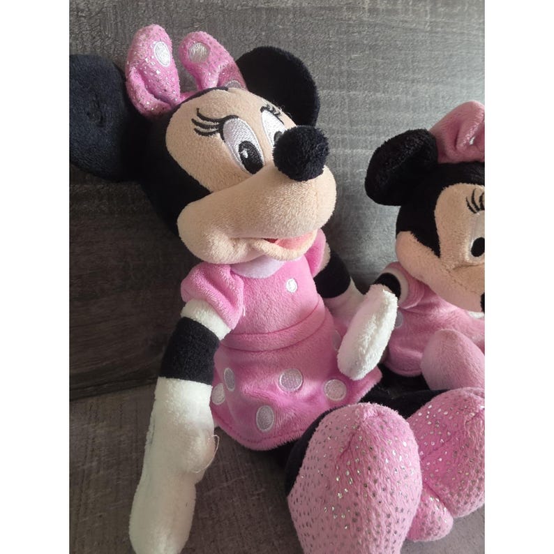 May include: Two Minnie Mouse plush toys. The plush toys have black ears, a beige face, and a black nose. They are wearing pink dresses with white polka dots and pink bows. The plush toys have pink sparkly shoes.
