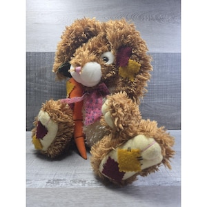 May include: A plush brown bunny rabbit with a fluffy texture, featuring a pink nose, white muzzle, and a carrot. The bunny has patches of yellow and burgundy on its paws and ears, and a pink floral scarf. The toy is designed for children.