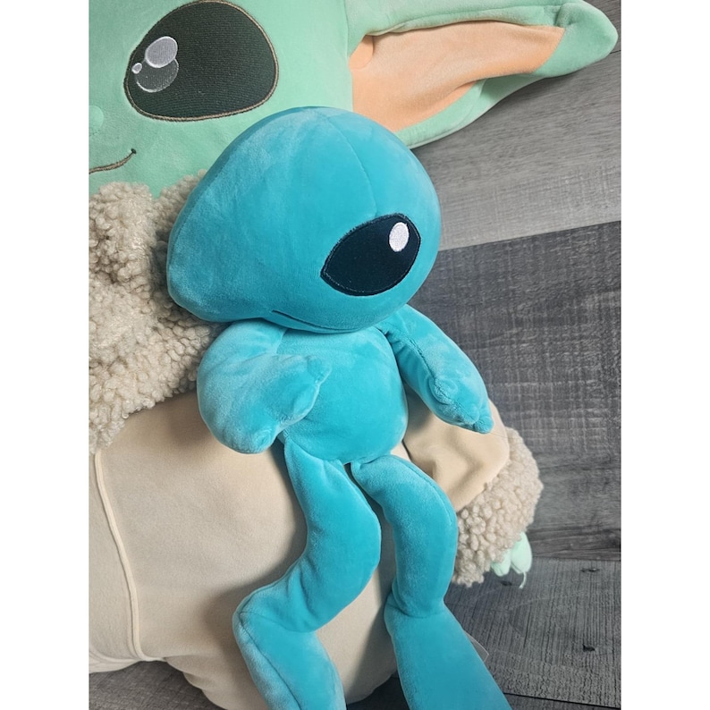 May include: A teal blue plush toy with a large eye and long legs. The toy is sitting on a plush, light brown and white toy.