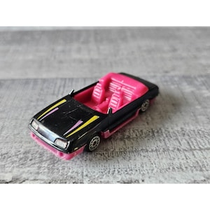 May include: Black toy car with pink interior and yellow accents. The car is a convertible with the top down.