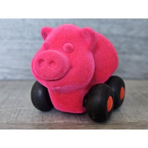 May include: A pink plush pig toy with black wheels. The pig has a smiling face and a pink nose.