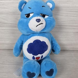 May include: A blue plush bear toy with a grumpy facial expression, featuring a white muzzle, a dark blue heart-shaped nose, and a white belly patch displaying a dark blue rain cloud with a small red heart and blue raindrops.