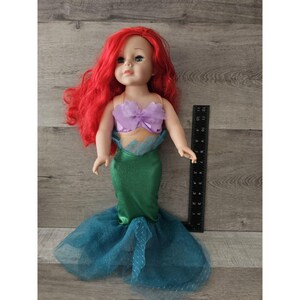 Puede incluir: Una mu&ntilde;eca de pelo rojo con un disfraz de sirena morado y verde. La mu&ntilde;eca mide aproximadamente 45 cm y tiene ojos azules. El disfraz incluye un top morado con un lazo y una cola verde con una falda de tul.