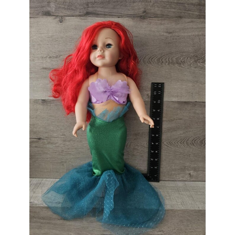 Puede incluir: Una mu&ntilde;eca de pelo rojo con un disfraz de sirena morado y verde. La mu&ntilde;eca mide aproximadamente 45 cm y tiene ojos azules. El disfraz incluye un top morado con un lazo y una cola verde con una falda de tul.