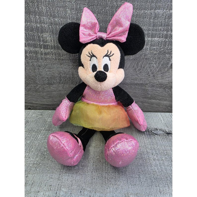 K&ouml;nnte beinhalten: Eine Minnie Mouse Pl&uuml;schpuppe mit rosa Schleife, Kleid und Schuhen. Die Puppe hat schwarze Ohren, Arme und Beine und ein cremefarbenes Gesicht mit gestickten Merkmalen. Das Kleid ist eine Kombination aus Rosa und Gelb.