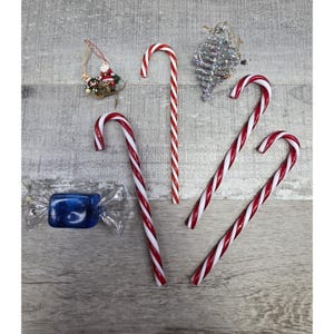 May include: An assortment of Christmas decorations on a gray wooden surface. The image features red and white striped candy canes, a blue wrapped candy, and silver and gold ornaments. The ornaments include a Santa Claus in a sleigh and a silver pinecone.