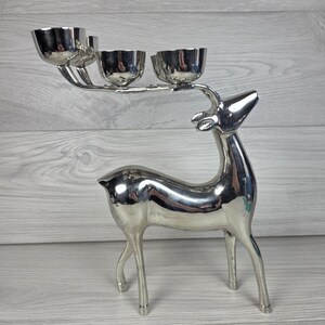 May include: A polished silver-coloured metal candle holder shaped like a standing deer. The deer's antlers extend horizontally to support three small, rounded, bowl-shaped candle cups arranged in a row along the top of the head.