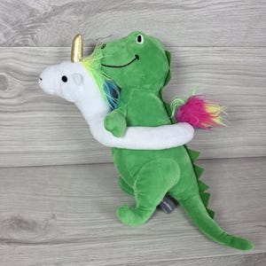 May include: A plush toy featuring a green dinosaur being hugged by a white unicorn. The unicorn has a gold horn and a rainbow-colored mane and tail. The dinosaur has a dark green back ridge and a smiling face. The plush is soft and textured.